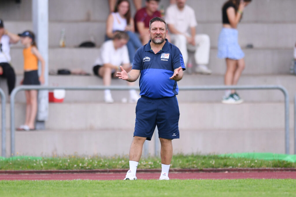 Mann in kurzer Hose und kurzem Shirt in blau und weiß an der Seitenlinie eines Fußballfelds.
